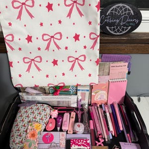 Peut inclure: Un sac fourre-tout blanc &agrave; motifs de n&oelig;uds et d'&eacute;toiles roses est suspendu au-dessus d'un plateau noir rempli de papeterie, dont des stylos roses, une liste de lecture 2026, un marque-page floral, un sachet de th&eacute; et un panneau noir Catching Dreams by Meg.
