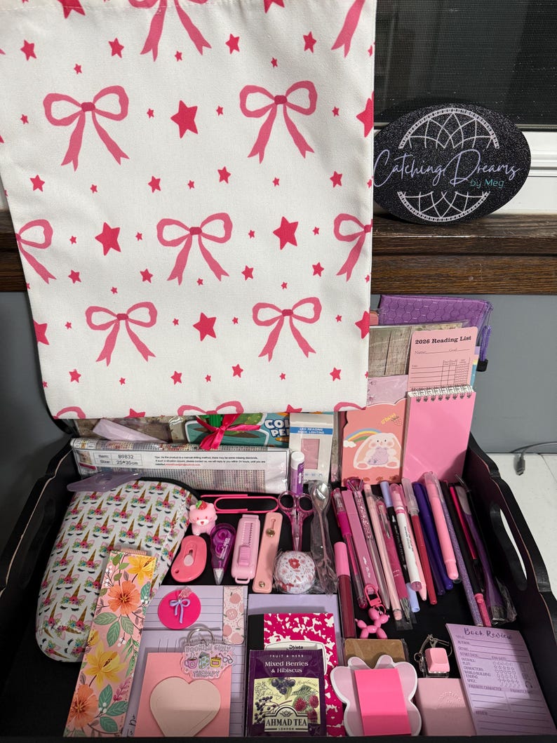 Peut inclure: Un sac fourre-tout blanc &agrave; motifs de n&oelig;uds et d'&eacute;toiles roses est suspendu au-dessus d'un plateau noir rempli de papeterie, dont des stylos roses, une liste de lecture 2026, un marque-page floral, un sachet de th&eacute; et un panneau noir Catching Dreams by Meg.
