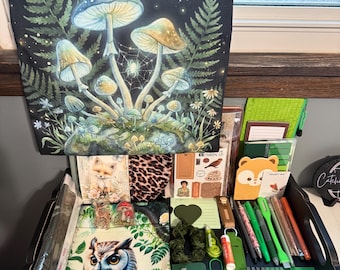 May include: A collection of stationery and craft items including a wall hanging with a moonlit mushroom forest, an owl-themed notebook, green pens, a mini composition book, a leopard print pouch, and various small accessories arranged on a tray.