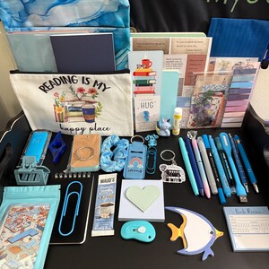 Peut inclure: Une collection d'articles de papeterie et d'accessoires de lecture comprenant une pochette en toile avec le texte Reading is my happy place, divers stylos bleus, un post-it en forme de c&oelig;ur, un bloc-notes en forme de poisson et des trombones.