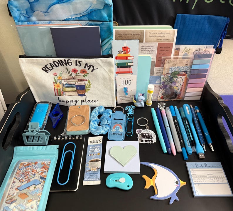 Peut inclure: Une collection d'articles de papeterie et d'accessoires de lecture comprenant une pochette en toile avec le texte Reading is my happy place, divers stylos bleus, un post-it en forme de c&oelig;ur, un bloc-notes en forme de poisson et des trombones.