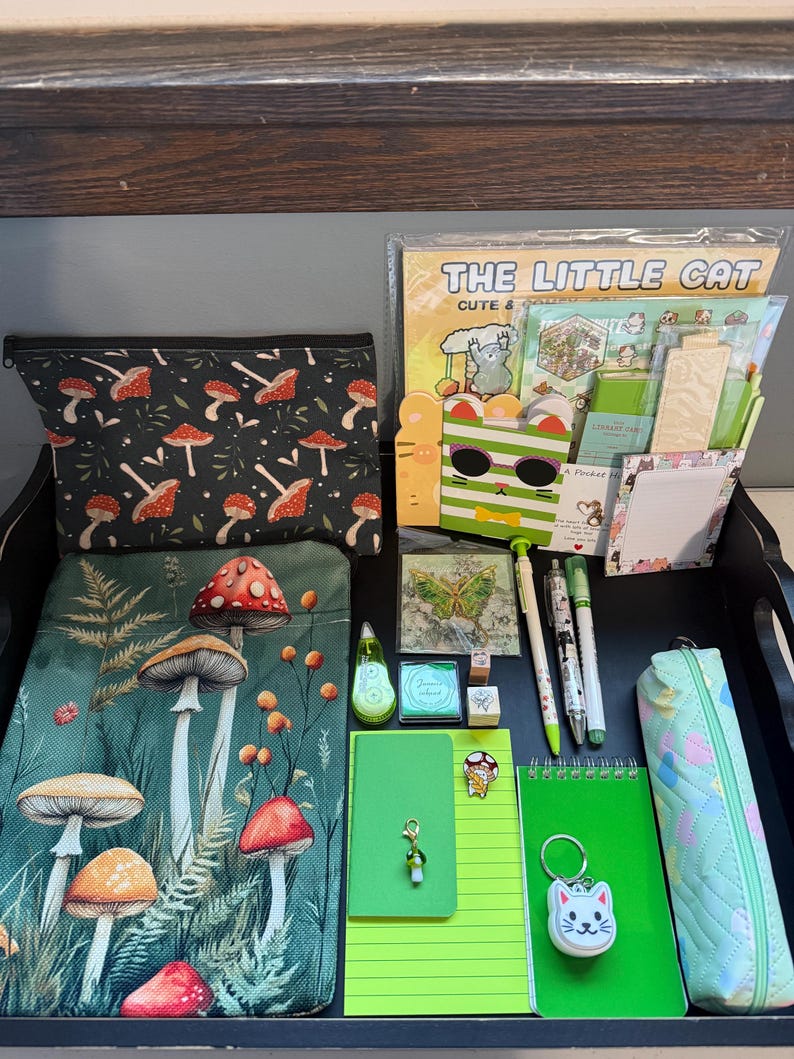 Peut inclure: Une collection d'articles de papeterie comprenant une pochette &agrave; motifs de champignons, un carnet vert, une trousse bleu clair, divers stylos, un porte-cl&eacute;s chat, un autocollant papillon et un bloc-notes intitul&eacute; The Little Cat avec des dessins.