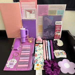 Peut inclure: Un ensemble de papeterie comprenant un carnet rose, un bloc-notes &agrave; spirales violet, un gobelet violet, un sachet de Maud's Raspberry Choco-Latte, des stylos, un marque-page floral, un chouchou violet, un post-it en fleur et divers autocollants.