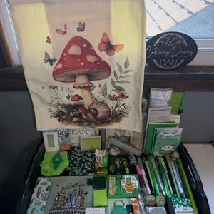 Peut inclure: Un sac fourre-tout en toile cr&egrave;me orn&eacute; d'un grand champignon rouge, d'un escargot et de papillons. En dessous se trouve une collection d'articles de papeterie verts, incluant des stylos, des carnets, un marque-page &agrave; motifs cactus et une pochette avec le texte "Just one MORE CHAPTER".