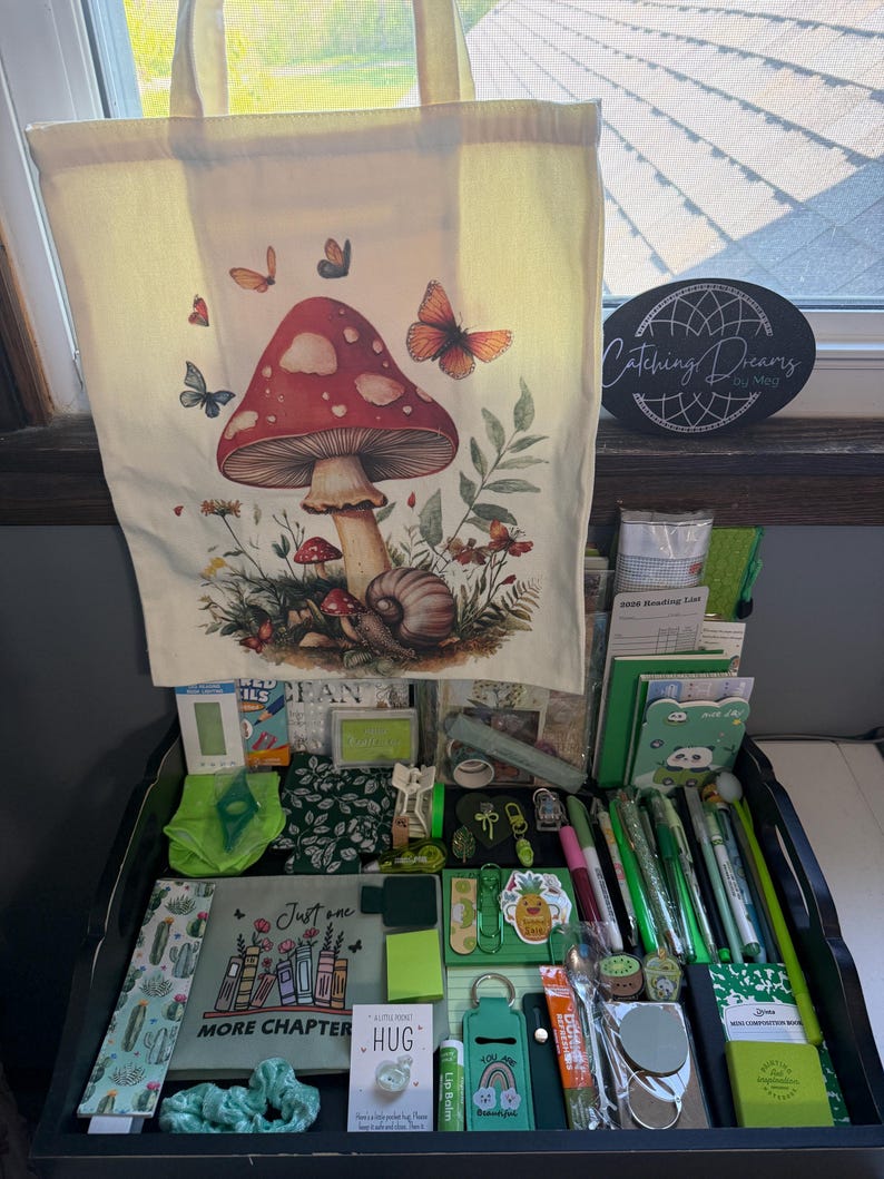 Peut inclure: Un sac fourre-tout en toile cr&egrave;me orn&eacute; d'un grand champignon rouge, d'un escargot et de papillons. En dessous se trouve une collection d'articles de papeterie verts, incluant des stylos, des carnets, un marque-page &agrave; motifs cactus et une pochette avec le texte "Just one MORE CHAPTER".