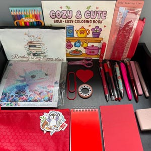 Peut inclure: Une collection de fournitures de papeterie et de loisirs cr&eacute;atifs comprenant un livre de coloriage intitul&eacute; Cozy & Cute, une trousse Reading is my happy place, un kit de broderie diamant axolotl, des crayons, des stylos rouges et un carnet.