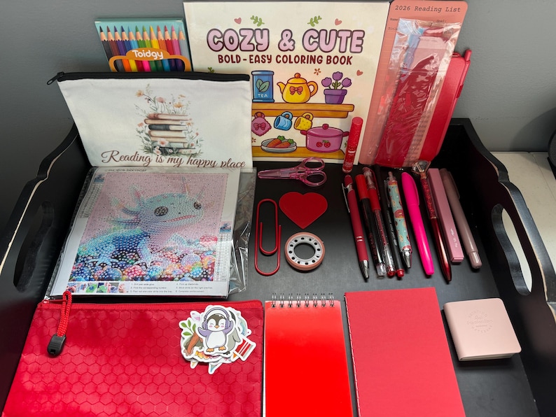 Peut inclure: Une collection de fournitures de papeterie et de loisirs cr&eacute;atifs comprenant un livre de coloriage intitul&eacute; Cozy & Cute, une trousse Reading is my happy place, un kit de broderie diamant axolotl, des crayons, des stylos rouges et un carnet.