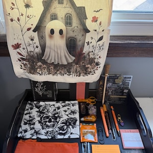 Peut inclure: Une tenture murale en tissu cr&egrave;me repr&eacute;sentant un fant&ocirc;me devant une maison, une pochette &agrave; motif de cr&acirc;ne noir et blanc, une pochette zipp&eacute;e orange, divers stylos, un petit balai, un sachet de th&eacute;, une cuill&egrave;re et divers articles de papeterie.