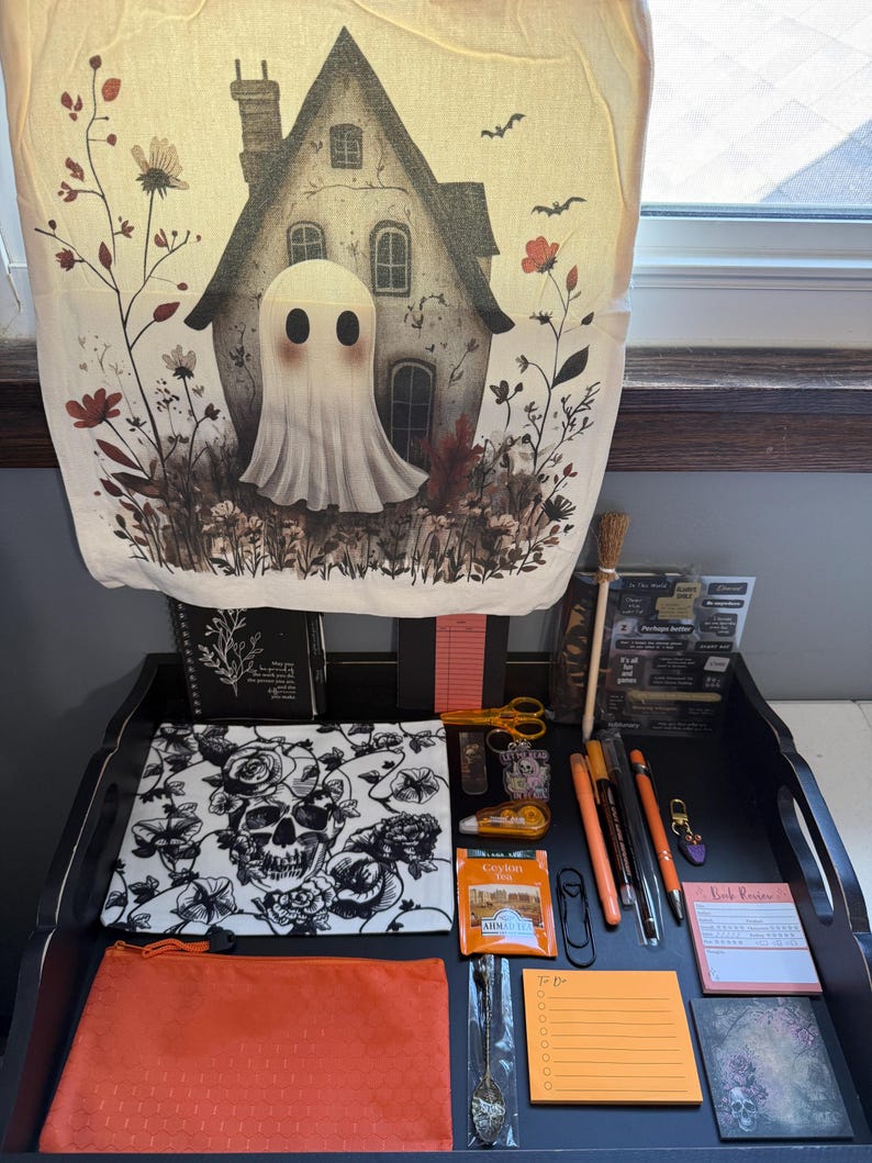 Peut inclure: Une tenture murale en tissu cr&egrave;me repr&eacute;sentant un fant&ocirc;me devant une maison, une pochette &agrave; motif de cr&acirc;ne noir et blanc, une pochette zipp&eacute;e orange, divers stylos, un petit balai, un sachet de th&eacute;, une cuill&egrave;re et divers articles de papeterie.