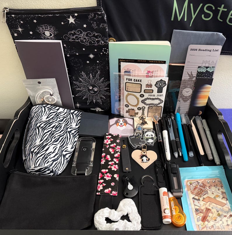 Peut inclure: Un plateau noir rempli de papeterie et d'accessoires, incluant une pochette zipp&eacute;e &agrave; motif c&eacute;leste, une housse &agrave; motif z&eacute;br&eacute;, une liste de lecture 2026, divers stylos, des autocollants, un porte-cl&eacute;s en forme de c&oelig;ur, un chouchou blanc et une petite pochette noire.