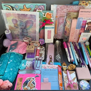 Peut inclure: Un ensemble de fournitures de papeterie et de loisirs cr&eacute;atifs comprenant un kit de broderie diamant avec un &eacute;l&eacute;phant, une liste de lecture 2026, divers stylos, des marque-pages, une lampe de lecture LED rose, une trousse turquoise et un bloc-notes.