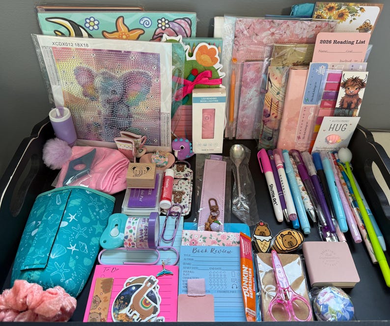 Peut inclure: Un ensemble de fournitures de papeterie et de loisirs cr&eacute;atifs comprenant un kit de broderie diamant avec un &eacute;l&eacute;phant, une liste de lecture 2026, divers stylos, des marque-pages, une lampe de lecture LED rose, une trousse turquoise et un bloc-notes.