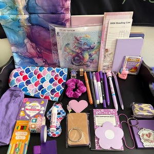 Peut inclure: Une collection d'articles de papeterie et de loisirs cr&eacute;atifs sur un plateau noir, comprenant un kit de peinture diamant dragon, une liste de lecture 2026, un carnet violet, une pochette &eacute;cailles de sir&egrave;ne, un mini livre de graffiti et des stylos.