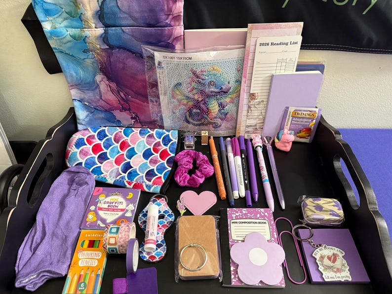 Peut inclure: Une collection d'articles de papeterie et de loisirs cr&eacute;atifs sur un plateau noir, comprenant un kit de peinture diamant dragon, une liste de lecture 2026, un carnet violet, une pochette &eacute;cailles de sir&egrave;ne, un mini livre de graffiti et des stylos.