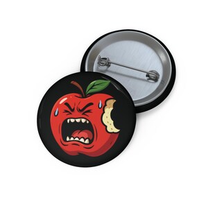 May include: A round, black button with a cartoon illustration of an angry, red apple with a bite taken out of it. The apple has a green leaf and is sweating. A second button is visible.
