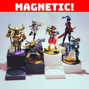 May include: A collection of seven character figurines, including a golden knight, Link, and others, displayed on tiered black and white platforms. The word "MAGNETIC!" is displayed in white text on a red background.