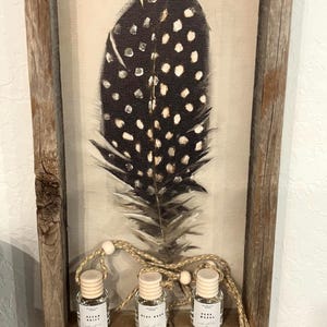 May include: A framed canvas print featuring a black and white feather design. Below the print, three small glass bottles with wooden lids are displayed, connected by a rope with wooden beads. The bottles have labels with text.