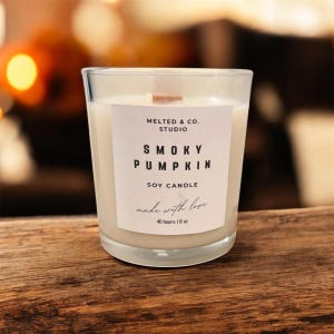 May include: A clear glass candle with a white label that reads "SMOKY PUMPKIN SOY CANDLE" from MELTED & CO. STUDIO. The candle has a wooden wick and is filled with a creamy wax. The label also states "made with love" and "40 hours / 8 oz".