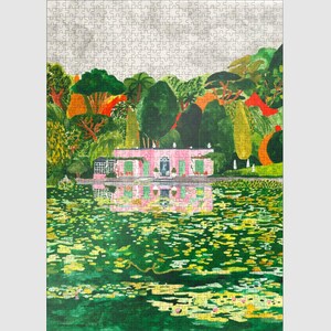 May include: A completed jigsaw puzzle featuring a pink house with green shutters reflected in a green pond filled with lily pads. The house is surrounded by lush green trees and foliage, with a cloudy sky background.