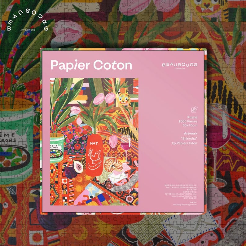 May include: A pink box of a 1000-piece puzzle with the title "Papier Coton" and the artwork "Shiracha" by Papier Coton. The puzzle image features a colorful still life with plants, food, and a tiger. The puzzle measures 50cm x 70cm.
