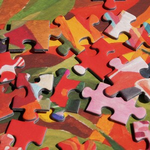May include: Close-up of a partially assembled jigsaw puzzle. The puzzle pieces are primarily red, with accents of pink, yellow, and white. The background features a colorful abstract design with green, orange, and brown hues. A fun activity for all ages.