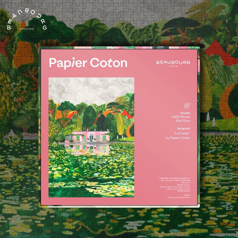 May include: A 1000-piece jigsaw puzzle with the artwork "La Folie" by Papier Coton. The puzzle box is pink and features a colorful painting of a pink building reflected in water, surrounded by lush green trees. The puzzle measures 50x70cm.