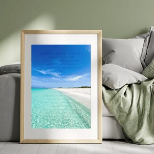 May include: A framed print featuring a beach scene with clear turquoise water, white sand, and a bright blue sky. The print includes text in a light blue font. The frame is a light wood color.