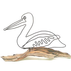 May include: A wire sculpture of a pelican featuring a continuous dark metal line forming the bird's silhouette, including a long beak and wing detail. The sculpture is mounted on a piece of natural, light brown driftwood base.