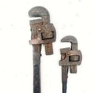 May include: Two heavy-duty metal pipe wrenches with a dark, weathered finish and visible signs of oxidation. Each tool features a serrated adjustable jaw, a threaded adjustment nut, and a long handle with a circular hole at the base for hanging.