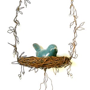 May include: A blue ceramic bird figurine sits inside a brown woven twig nest. The nest is suspended by a thin, twisted bronze-colored wire frame that forms a loop above and features decorative wire tendrils hanging below the nest structure.