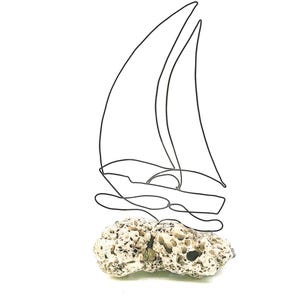 May include: A minimalist wire sculpture of a sailboat with two sails, atop two textured, light-colored rocks. The black wire art piece is set against a white background, creating a contrast.