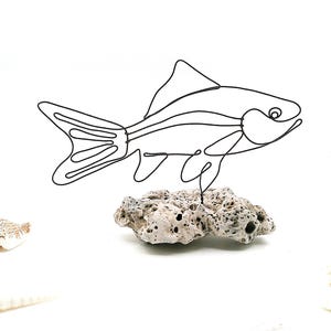 May include: A black wire sculpture of a fish, mounted on a piece of textured, light gray rock. The fish has a streamlined body, fins, and a detailed tail. The sculpture is set against a white background, with seashells in the corners.