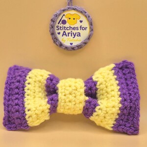 May include: A handmade crochet bow tie in purple and yellow stripes. The bow tie is centered in the image, with a circular tag above it that reads "Stitches for Ariya by Natasha."