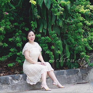 May include: A woman wearing a white crocheted dress with a scalloped hem and short sleeves. She is sitting on a stone wall in front of a lush green background.