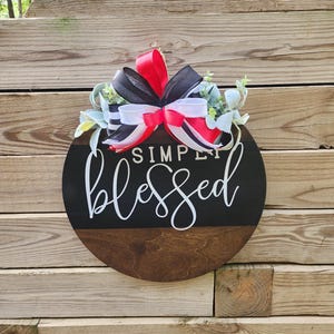 Puede incluir: Un letrero redondo de madera con las palabras "SIMPLY blessed" en escritura blanca. La mitad superior es negra, la inferior está teñida de marrón. Un lazo decorativo con cintas rojas, negras y blancas y vegetación se encuentra en la parte superior.