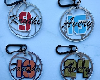 May include: Four clear acrylic keychains with black carabiner clips. Each keychain features a name and number in various colors: red 9, blue 15, orange 13, and gold 24. The names are in black script, and the numbers are in a block font.