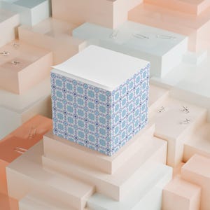 May include: A square notepad with a white top and patterned sides. The sides feature a repeating geometric design in light blue and purple on a white background. The notepad is displayed on a multi-level, pastel-colored platform with scattered paper clips.