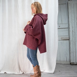 May include: A burgundy hooded sweater with a long, asymmetrical hemline. The sweater has a large pocket on the front and is made of a soft, textured fabric.