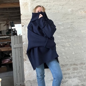 May include: A navy blue oversized hooded tunic with wide sleeves and a high collar. The tunic is paired with light blue denim jeans. The background is a textured, light-colored stone wall.