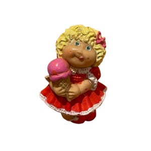 May include: A vintage Cabbage Patch Kids figurine of a girl holding a pink ice cream cone. The doll has blonde curly hair, blue eyes, and is wearing a red dress with white trim. A pink bow adorns her hair.