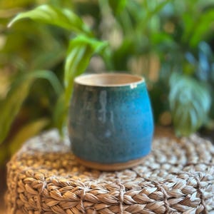 May include: A ceramic tumbler with a rounded silhouette, featuring a glossy glaze that transitions from a deep teal at the base to a lighter, mottled blue near the rim, with a smooth, unglazed clay bottom edge visible at the very base.