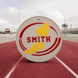 May include: A round, light gray sign with the name "SMITH" in red, stylized text. The sign features a yellow winged emblem and a yellow torch design, set against a red curved border. The sign is on a red track.