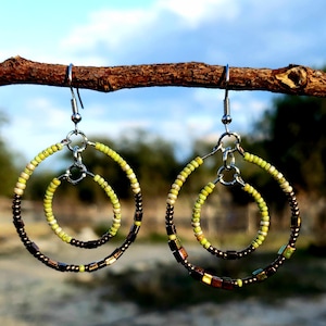 May include: Handmade beaded hoop earrings. The earrings feature a double hoop design with a smaller inner hoop. The outer hoops are dark brown with square beads, while the inner hoops are light green. The earrings hang from silver-colored hooks.