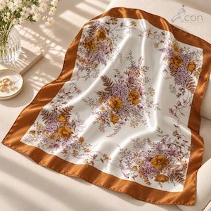 May include: A square satin scarf featuring a white centre with a floral pattern in shades of gold, brown, and purple, surrounded by a solid brown border. The fabric has a smooth, reflective finish and is draped to show its soft, flowing texture.