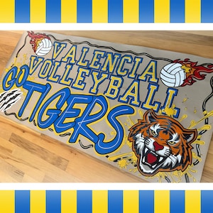 May include: A banner with the words "Valencia Volleyball Tigers" in blue and yellow. The banner features a tiger head graphic and volleyballs with flames. The banner is on a light brown background.