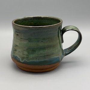 May include: A handmade ceramic mug with a rounded body and a sturdy handle. The mug features a blend of green and blue glaze over a brown base, creating a textured, earthy appearance. The mug is designed for hot beverages.