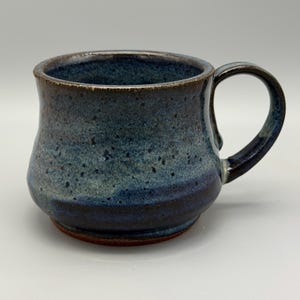 May include: A handmade ceramic mug with a dark blue and teal glaze. The mug has a rounded body, a sturdy handle, and a dark brown rim. The base of the mug is a reddish-brown color.