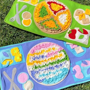 May include: Two colorful felt food play mats on green grass. One mat is green with a felt plate of food in yellow, red, green, and white. The other mat is blue with a felt plate of food in pink, yellow, green, blue, and white.