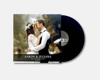 May include: A black vinyl record next to a wedding album. The album cover features a photo of a couple embracing, with the names "Aaron & Juliana" and the date "05-05-2025" printed on the bottom.