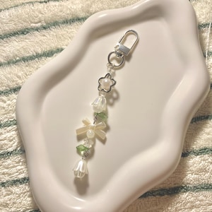 May include: A decorative keychain with a silver clasp and a charm made of white flower beads, small green beads, and cream bows. The keychain is presented on a white, wavy-edged tray.
